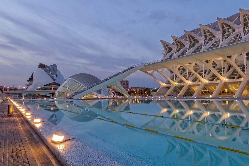 Edificios modernos e iluminados con arquitectura futurista reflejados en una larga piscina de agua al anochecer bajo un cielo nublado.