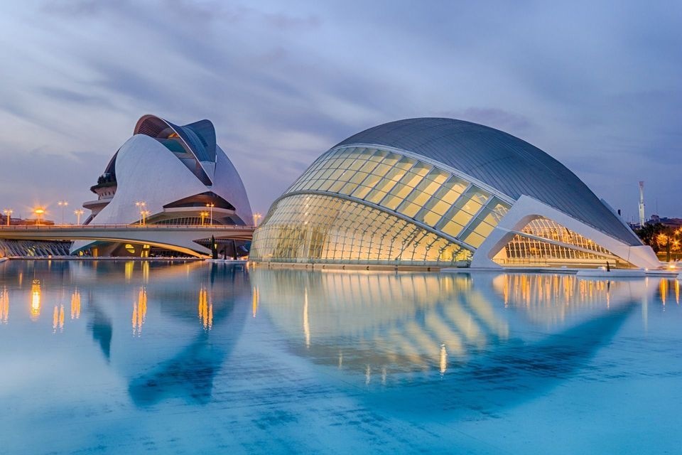 Dos edificios de arquitectura moderna iluminados al atardecer, sus luces reflejándose en una tranquila piscina de agua.