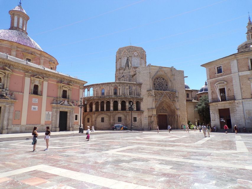 Una plaza pública amplia y soleada con adoquines estampados, rodeada de grandes edificios históricos bajo un cielo azul claro.