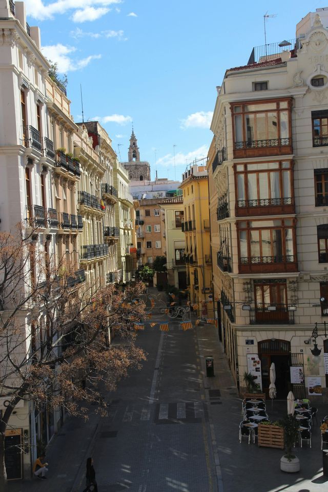 Una vista desde lo alto de una calle europea estrecha, flanqueada por edificios tradicionales y un campanario distante bajo un cielo azul.