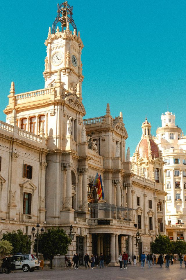 Un edificio de piedra ornamentado con una gran torre del reloj se alza contra un cielo azul claro, con gente caminando en la plaza de abajo.