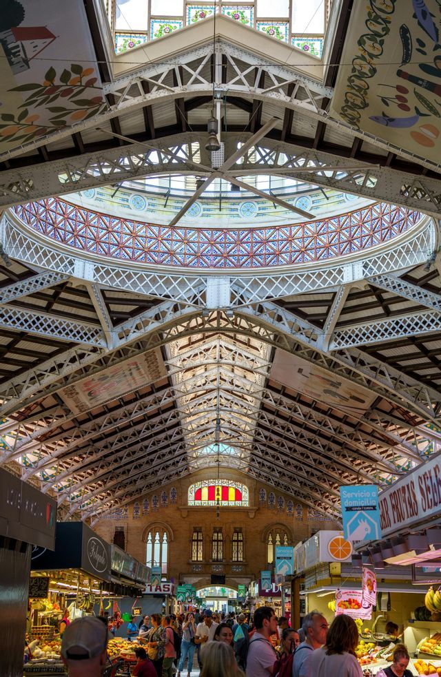 Un mercado interior concurrido con un techo alto y ornamentado que presenta una gran cúpula decorada y cerchas de metal blancas.