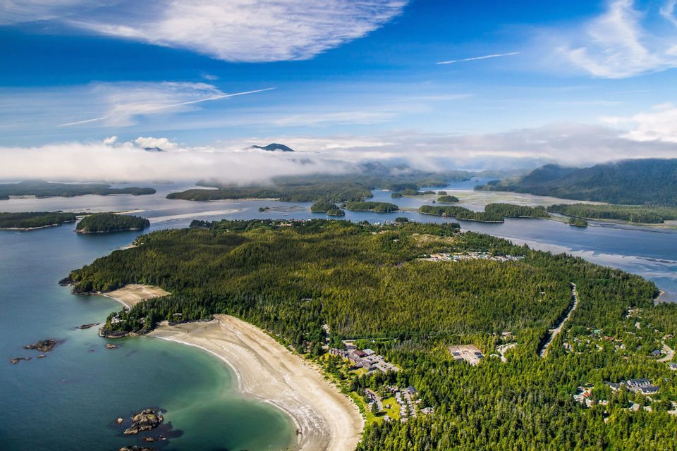 Vue aérienne d'un archipel boisé avec une plage de sable incurvée, entouré d'eau et de petites îles sous un ciel partiellement nuageux.