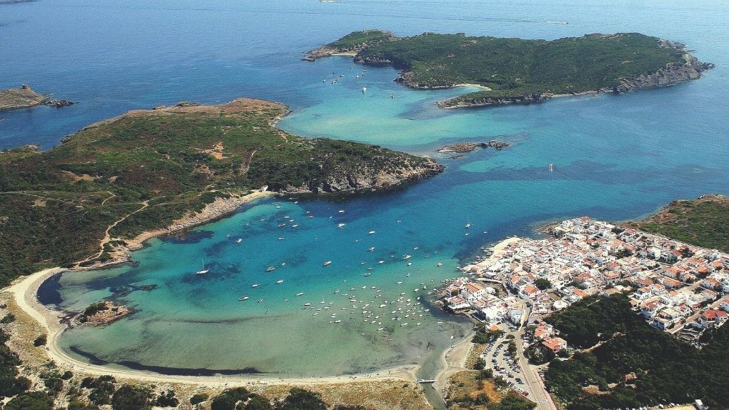 An aerial view of a coastal town with white buildings next to a turquoise bay filled with anchored boats.