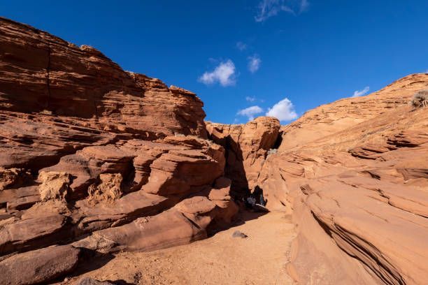 Dos personas de un viaje en grupo de WeRoad descansan a la sombra de un estrecho sendero de cañón de arenisca bajo un cielo azul brillante.