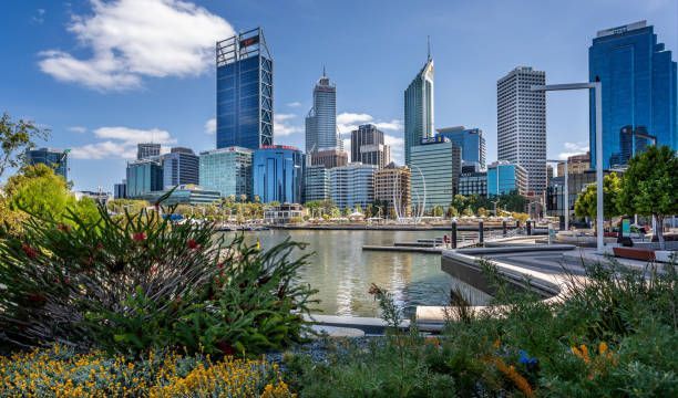 Un parque frente al mar con arbustos verdes y flores que da a un moderno horizonte urbano con rascacielos bajo un cielo parcialmente nublado.