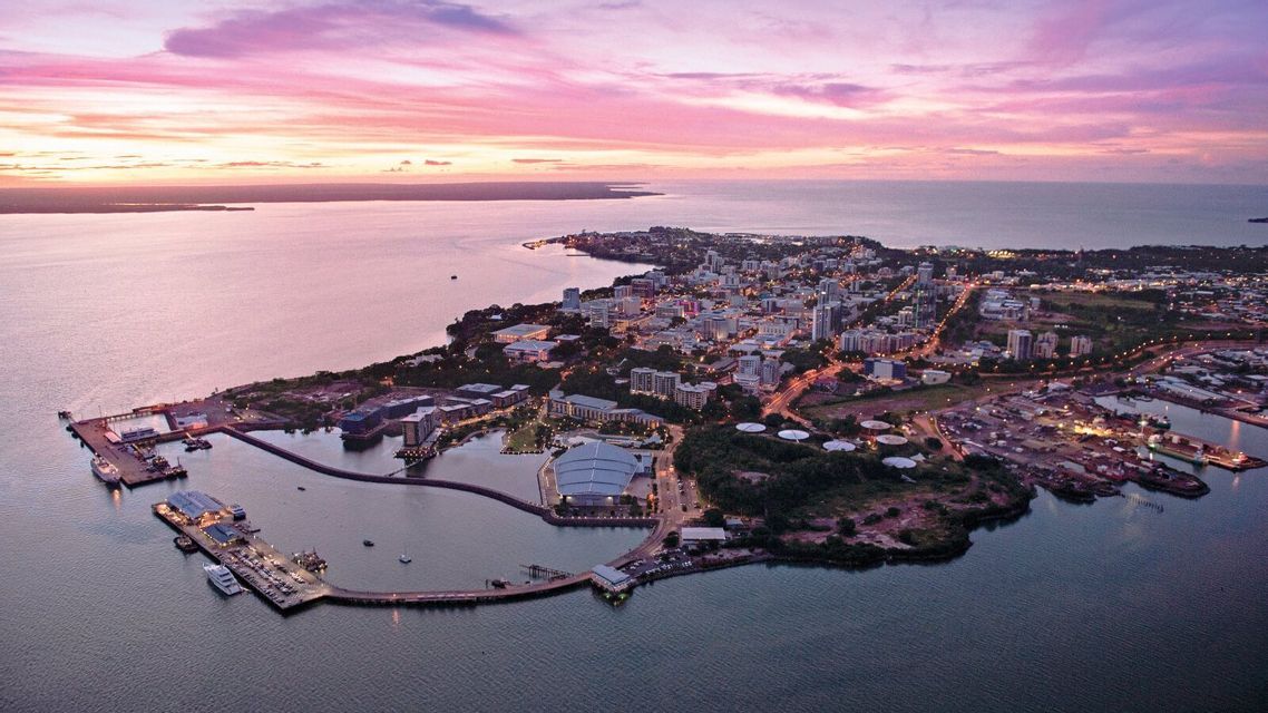 Una vista aérea de una ciudad costera con un gran puerto, iluminada por los tonos rosados y morados del atardecer.