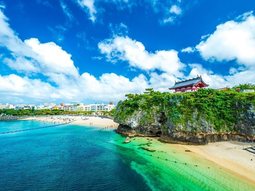 Un santuario giapponese con un tetto rosso si erge su una scogliera rocciosa e verde sopra una spiaggia sabbiosa con acqua turchese sotto un cielo azzurro.