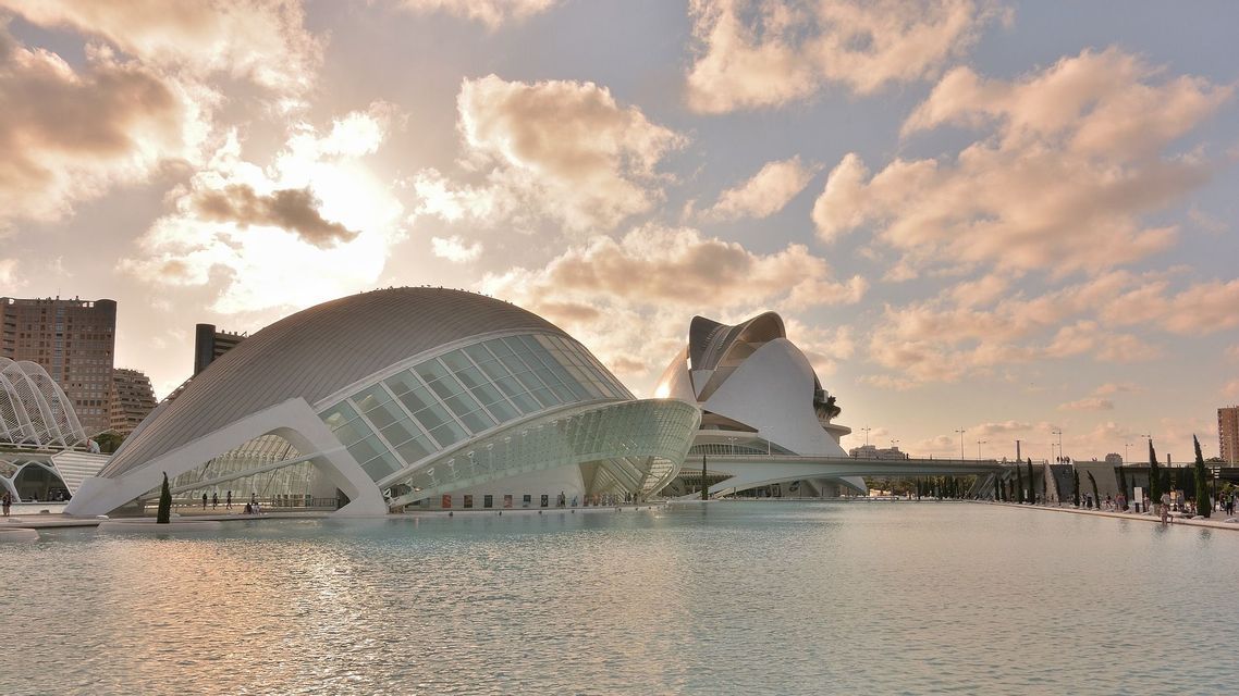 Dos grandes edificios blancos modernistas con fachadas de cristal situados junto a una gran lámina de agua bajo un cielo nublado al atardecer.