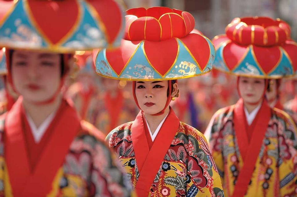 Una persona con trucco tradizionale indossa un costume colorato e un cappello rosso e blu ornato durante una parata culturale.