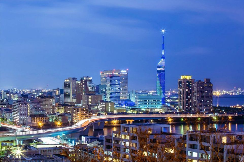 Uno skyline cittadino illuminato al tramonto, con un'imponente torre blu e un ponte con semafori che attraversa uno specchio d'acqua.