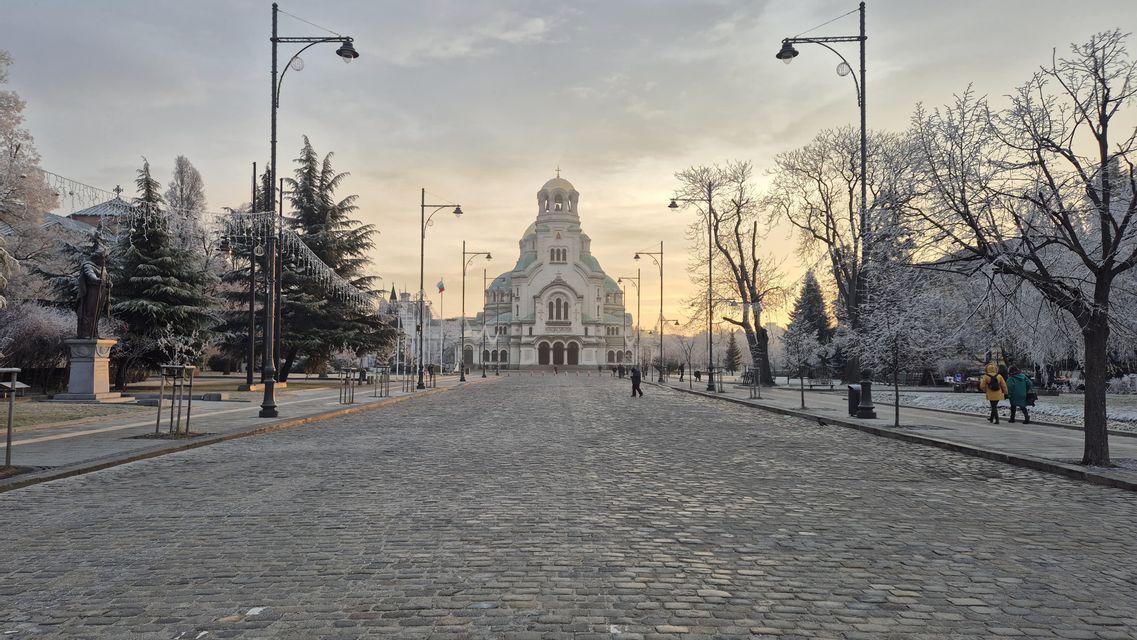 Un'ampia strada acciottolata conduce verso una grande cattedrale a cupola, affiancata da alberi e lampioni coperti di brina all'alba.
