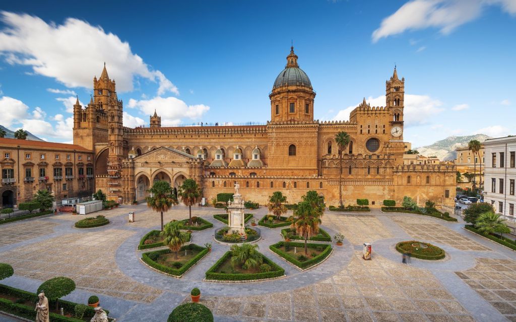 Una grande cattedrale color sabbia con un'ampia cupola si erge dietro una piazza ben curata con giardini e palme sotto un cielo blu.