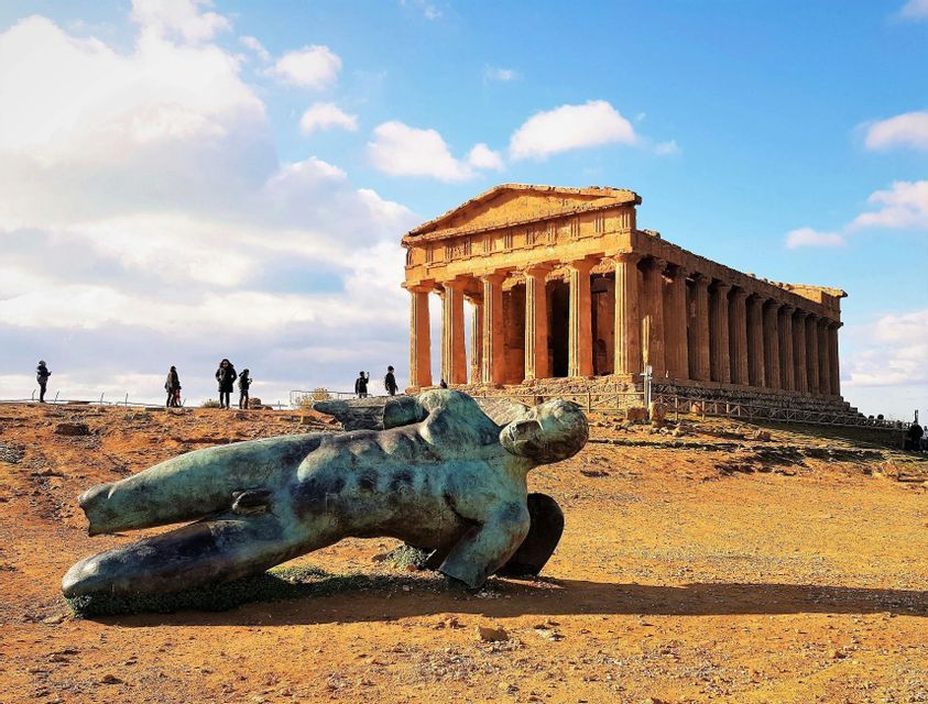 Una grande statua di bronzo giace caduta su una collina polverosa, con un antico tempio e alcune persone sullo sfondo.