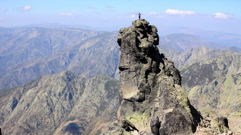 Una persona se alza con los brazos extendidos en la cima de un escarpado pico rocoso, contemplando una vasta cordillera.