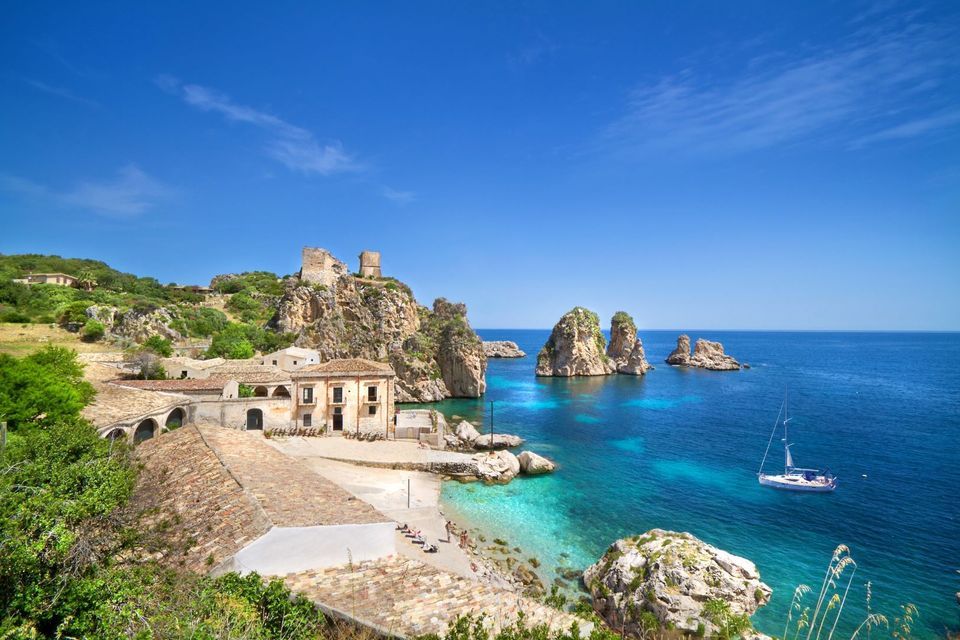 Un borgo costiero con le rovine di un castello su una scogliera, che si affaccia su una baia turchese con una barca a vela e grandi formazioni rocciose.