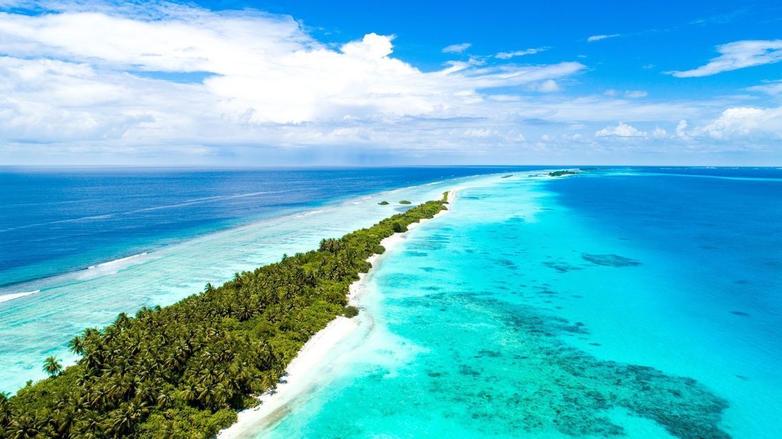 Veduta aerea di una stretta isola tropicale con palme e una spiaggia di sabbia bianca, che separa l'acqua turchese dall'oceano blu profondo.