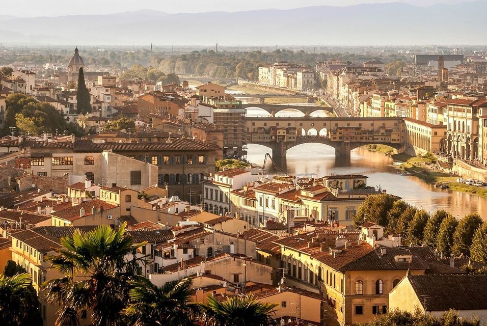An aerial view of a historic city with terracotta roofs and a river crossed by several arched bridges, bathed in golden light.