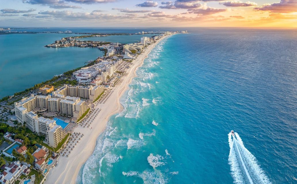 An aerial view of a hotel-lined coastline with a white sand beach, turquoise waters, and a motorboat at sunset.