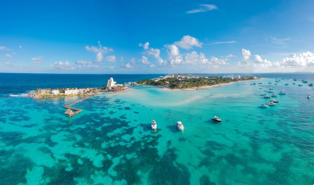 Aerial view of a tropical peninsula with resorts and boats anchored in clear, turquoise water under a blue sky with clouds.