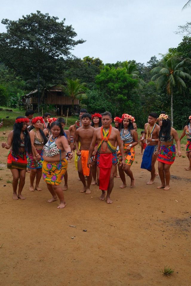 Un gruppo di indigeni con abiti tradizionali colorati e corone di fiori danzano a piedi nudi su una radura sterrata in un villaggio nella giungla.