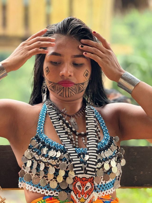 Una donna con trucco geometrico sul viso guarda in basso mentre indossa abiti tradizionali con perline adornati con monete.