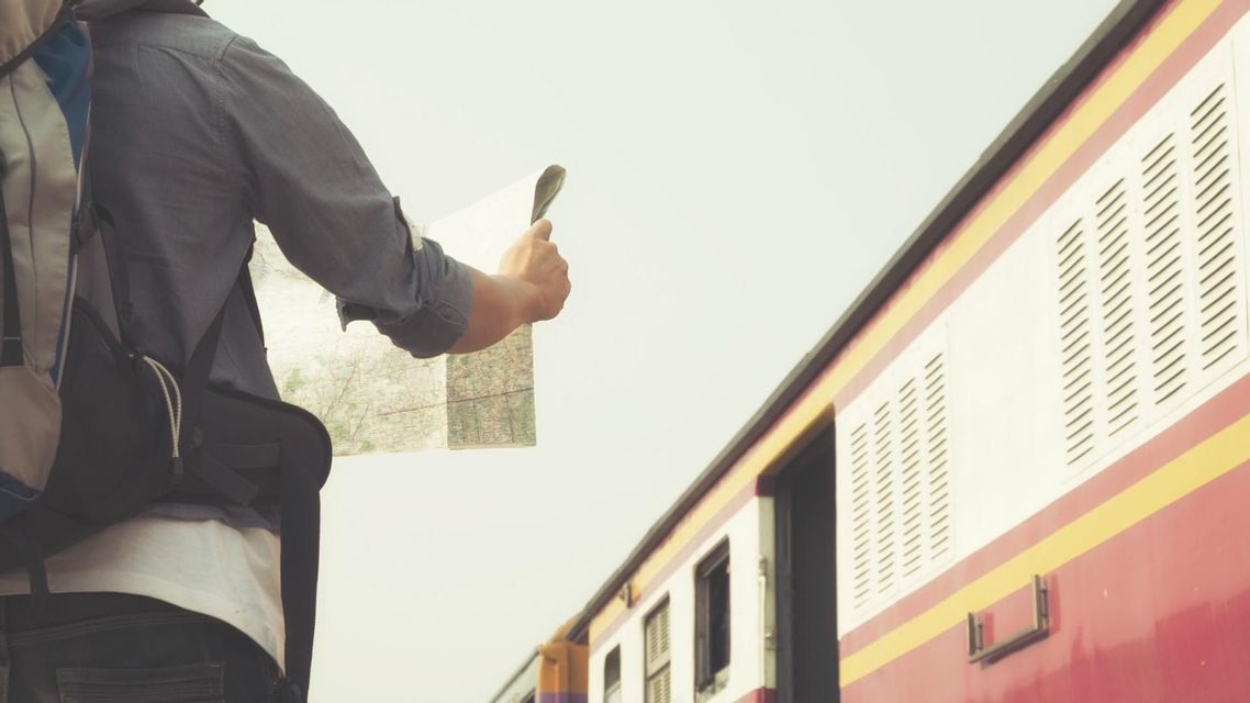 Un viaggiatore con uno zaino che tiene una mappa di carta si trova accanto a un treno rosso e giallo.