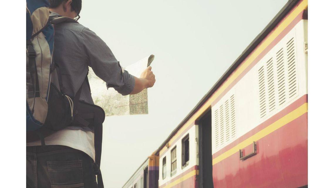Un viaggiatore con uno zaino sta su una piattaforma, studiando una mappa accanto a un treno fermo.