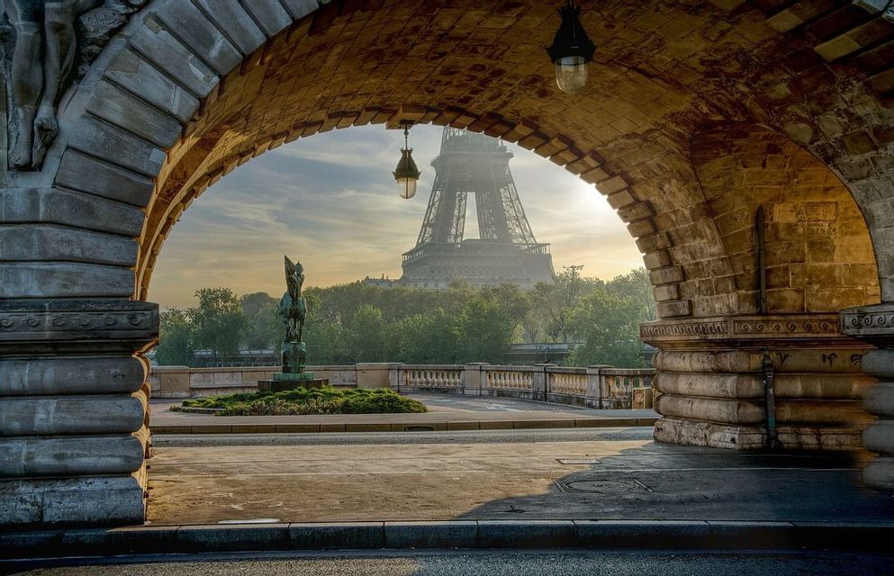 La Torre Eiffel incorniciata dall'arco in pietra di un ponte all'alba, con una statua equestre in bronzo visibile in primo piano.