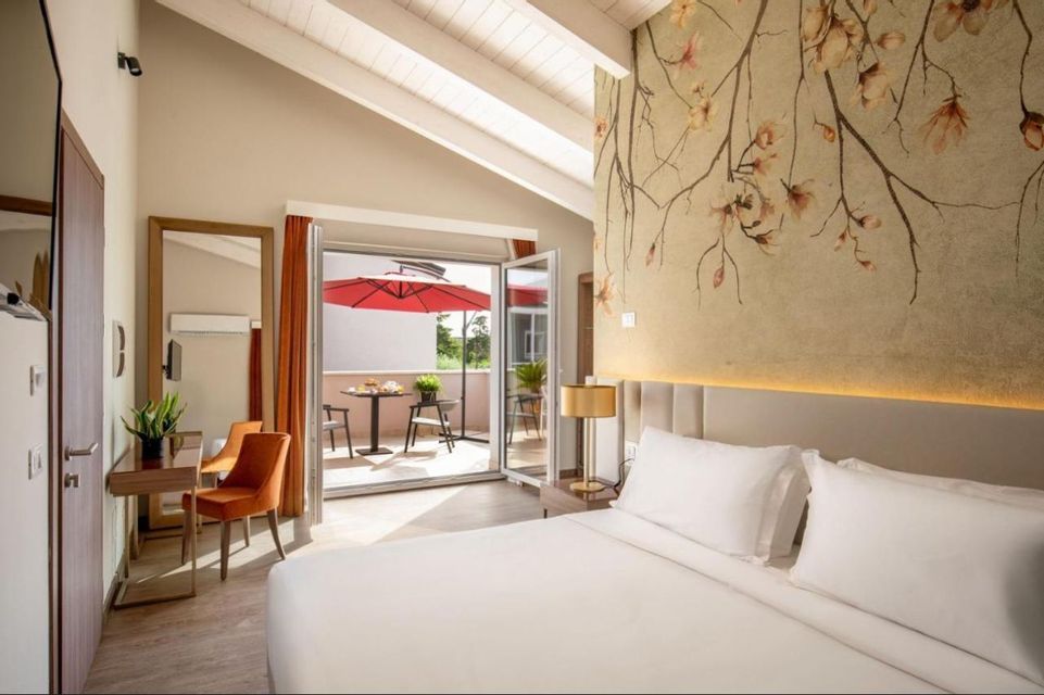 Una camera d'albergo con un letto bianco, una parete decorativa floreale e porte a vetro che si aprono su un balcone con un ombrellone rosso.