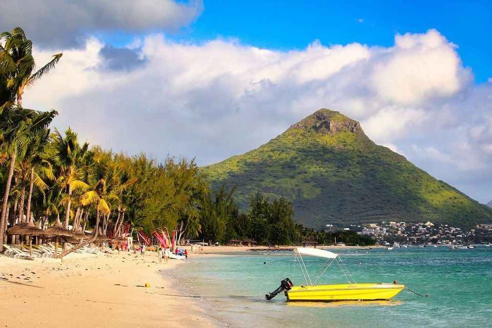 Un bateau à moteur jaune amarré dans les eaux turquoise d'une plage de sable, avec une grande montagne verte se dressant en arrière-plan sous un ciel bleu.