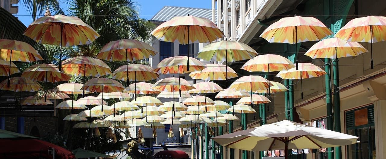 Des dizaines de parapluies ouverts avec un motif floral jaune et orange sont suspendus au-dessus d'une rue de ville sous un ciel bleu.