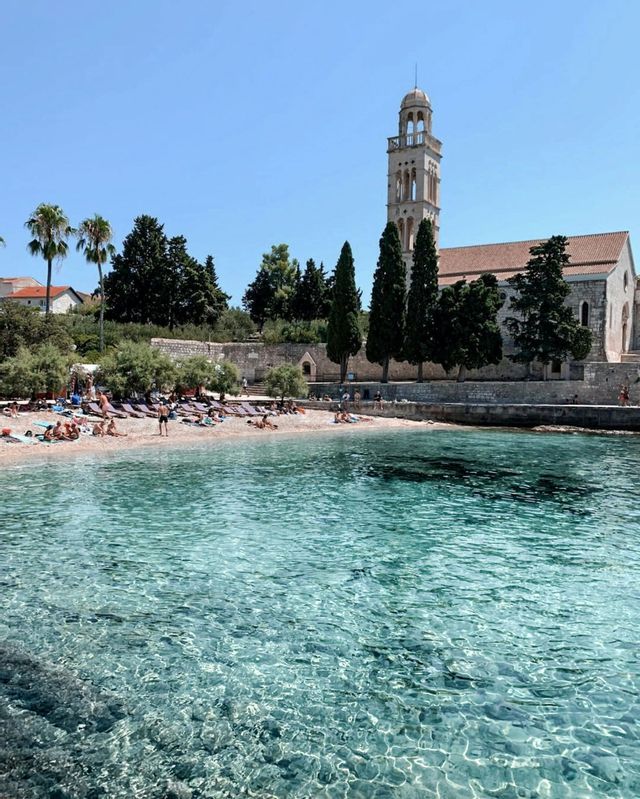 Persone che prendono il sole su una spiaggia sabbiosa accanto a un'acqua limpida e turchese, con una grande chiesa in pietra e un campanile sullo sfondo.