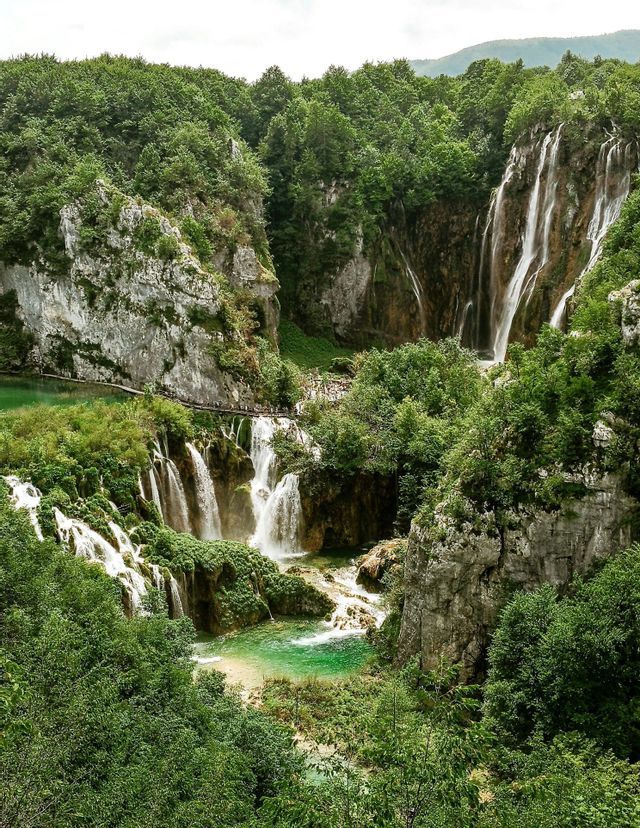 Una serie di cascate scende lungo formazioni rocciose muschiose in una pozza turchese, circondata da una fitta foresta verde e da una passerella con persone.