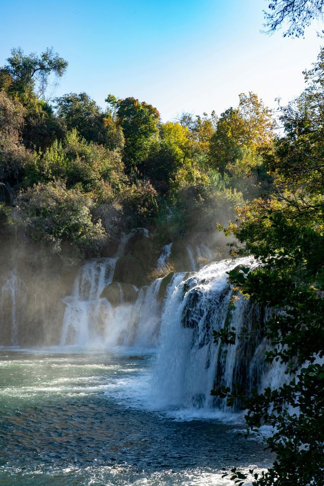 Una vasta cascata si getta in un fiume, creando nebbia alla luce del sole. È circondata da una fitta foresta sotto un cielo azzurro e limpido.
