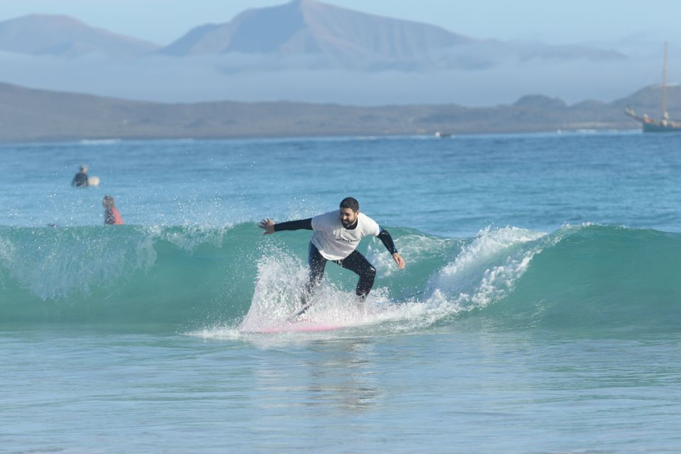 Ein Mann in einem Neoprenanzug und weißem T-Shirt surft auf einer türkisfarbenen Welle auf einem pinken Surfbrett, mit fernen Bergen im Hintergrund.
