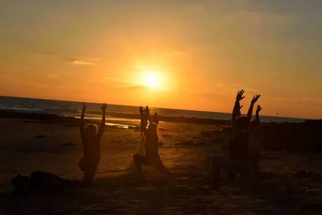 Silhouetten einer WeRoad-Gruppenreise beim Yoga am Strand mit erhobenen Armen während eines goldenen Sonnenuntergangs.