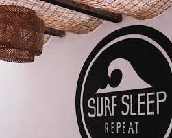 Ein schwarzes rundes Schild an einer weißen Wand mit den Worten 'SURF SLEEP REPEAT' und einer stilisierten Wellengrafik.