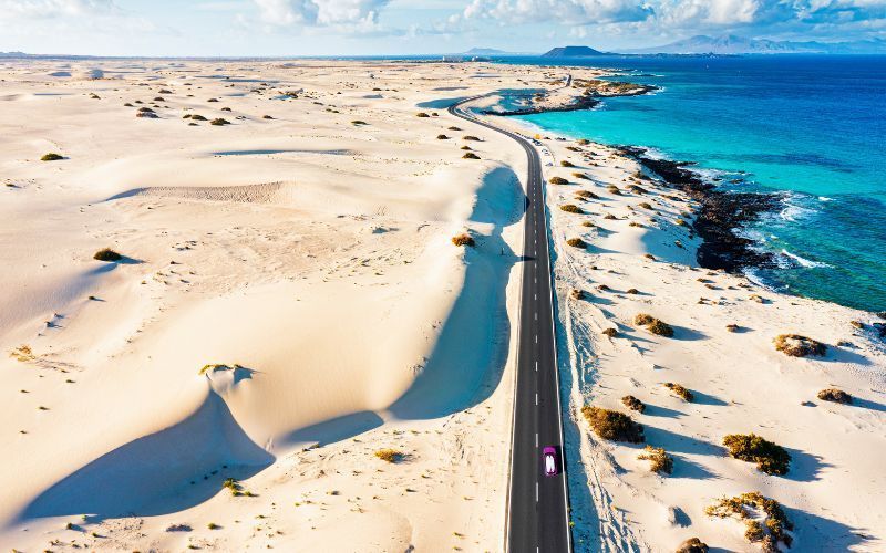 Luftaufnahme einer Küstenstraße zwischen weiten weißen Sanddünen und einem türkisfarbenen Meer, mit einem einzelnen Auto darauf.