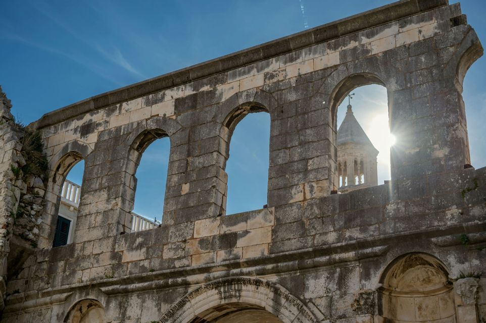 Una vista en ángulo bajo de una ruina de piedra desgastada con aberturas arqueadas, con un campanario visible contra un cielo azul soleado.
