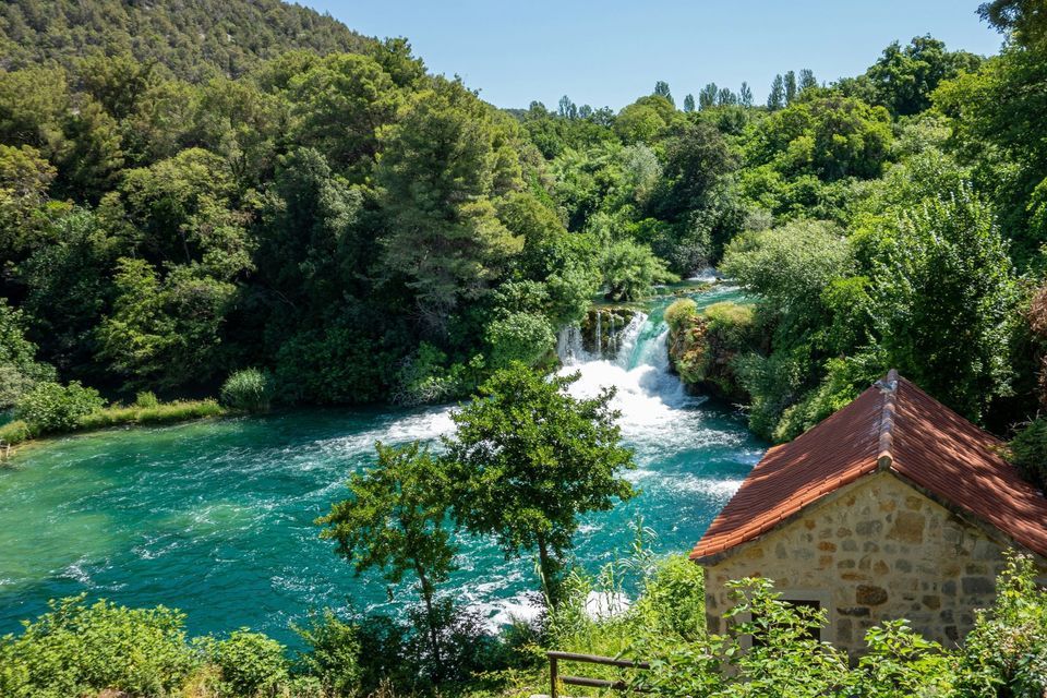 Une maison en pierre au toit de tuiles rouges surplombe une rivière turquoise et des cascades s'écoulant à travers une forêt verdoyante et luxuriante.