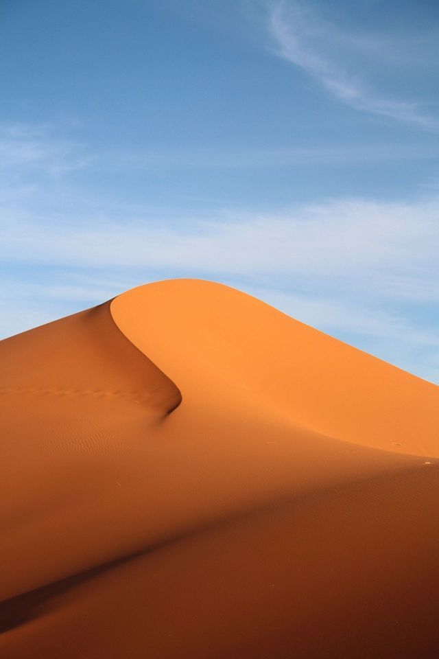 La cresta afilada y curvada de una duna de arena naranja, marcada con huellas, destaca sobre un cielo azul con nubes claras.