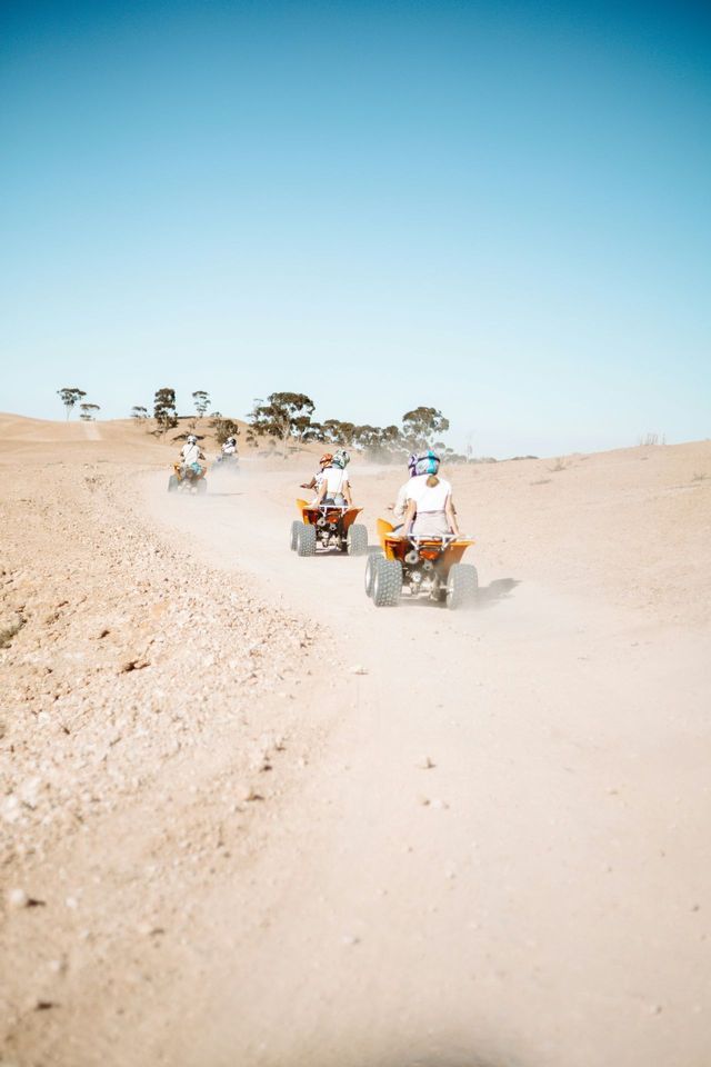 Un viaje en grupo de WeRoad en quads recorre en fila un camino de tierra polvoriento en un paisaje árido con árboles distantes.
