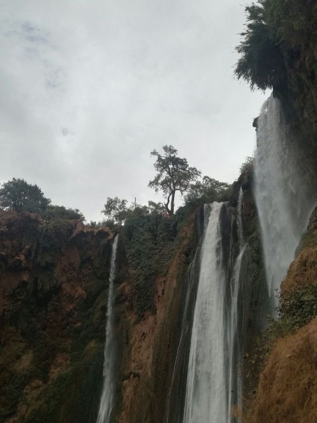 Múltiples cascadas descienden por un acantilado rocoso y escarpado cubierto de vegetación verde bajo un cielo nublado.