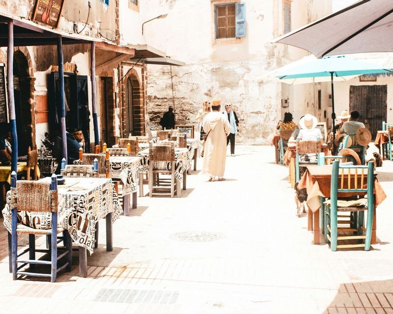 Mesas y sillas de una cafetería al aire libre flanquean una calle estrecha y soleada donde la gente camina y come.