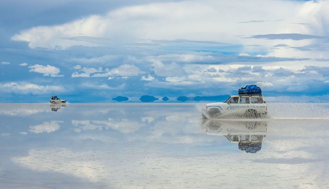Two white off-road vehicles drive through a vast, shallow body of water that perfectly reflects the cloudy sky.