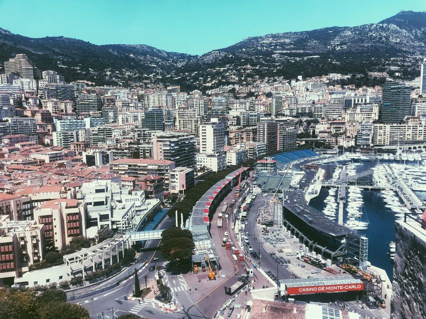 Veduta diurna dall'alto di Monte Carlo, che mostra fitti edifici cittadini, un porto turistico ricolmo di yacht e le montagne sullo sfondo.