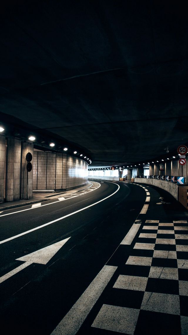 Una strada vuota si snoda attraverso un tunnel buio e illuminato con frecce bianche e un motivo a scacchiera dipinti sull'asfalto.