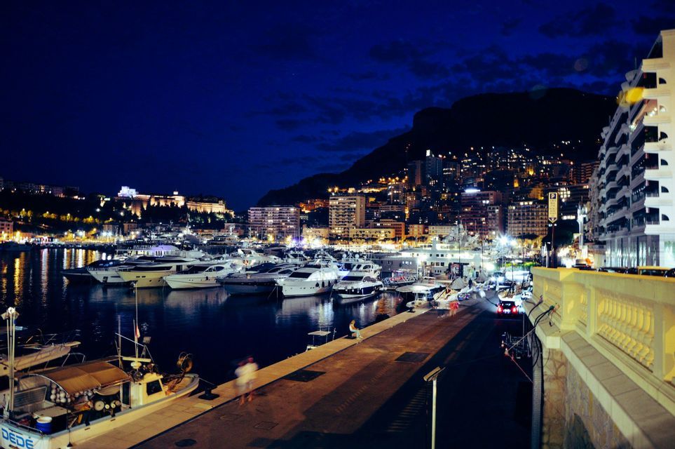 Yacht ormeggiati in un porto di notte, con uno skyline illuminato su una collina sullo sfondo.