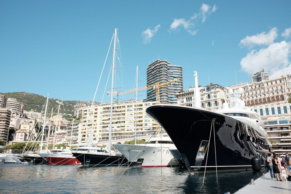 Un grande yacht nero e altre imbarcazioni sono ormeggiati in un porto di fronte a una collina densamente edificata sotto un cielo azzurro.